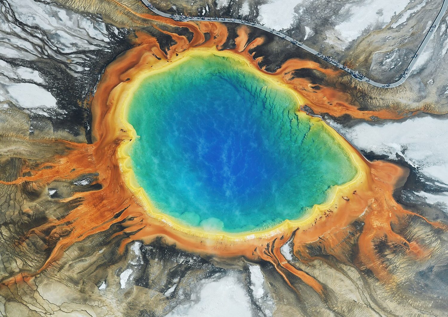 An aerial view of a hot spring: a deep blue centre ringed by bands of green, yellow and orange spreading out across the ground