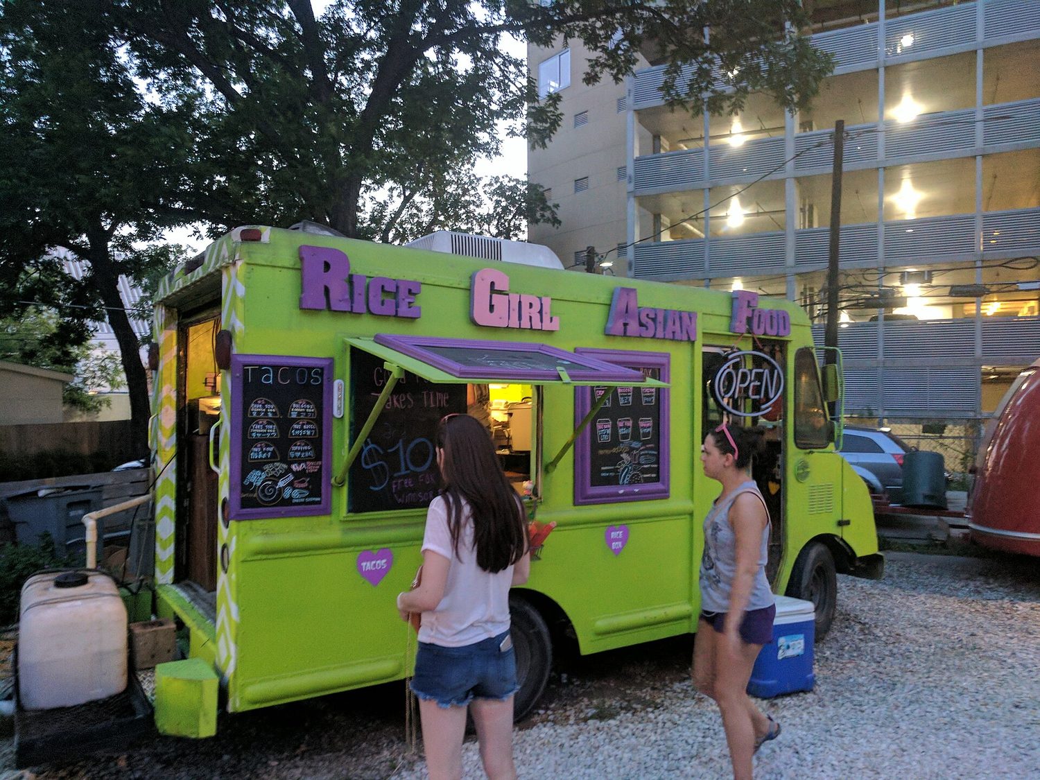 A brightly painted street-food truck with an "OPEN" sign and a menu, with two customers ordering at the window