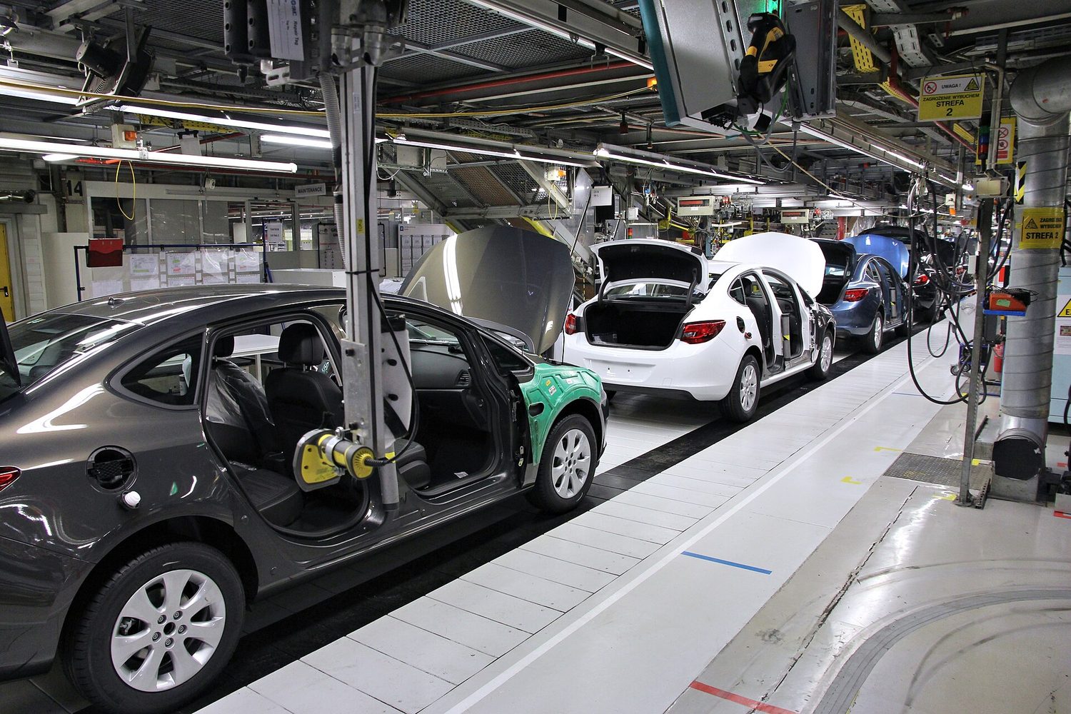 A car assembly line inside a factory: part-built cars in sequence along a moving line, with overhead tooling