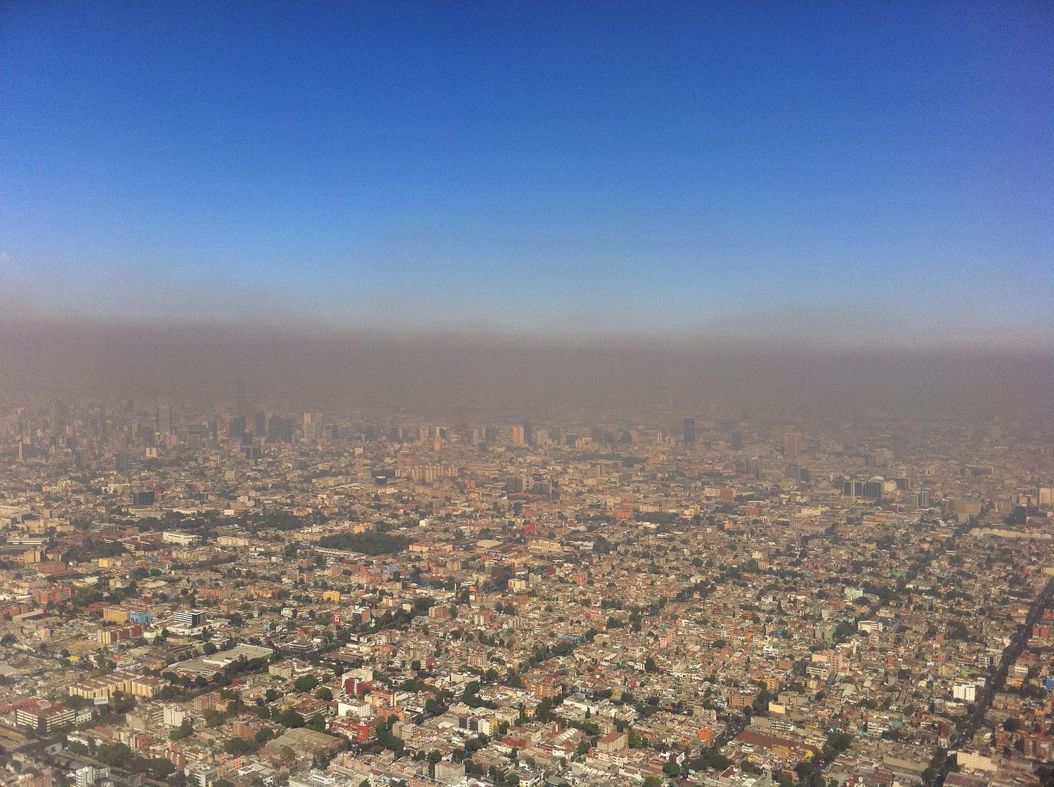 An aerial view of a large city with a distinct brown-grey layer of haze sitting over the buildings under a clear blue sky above