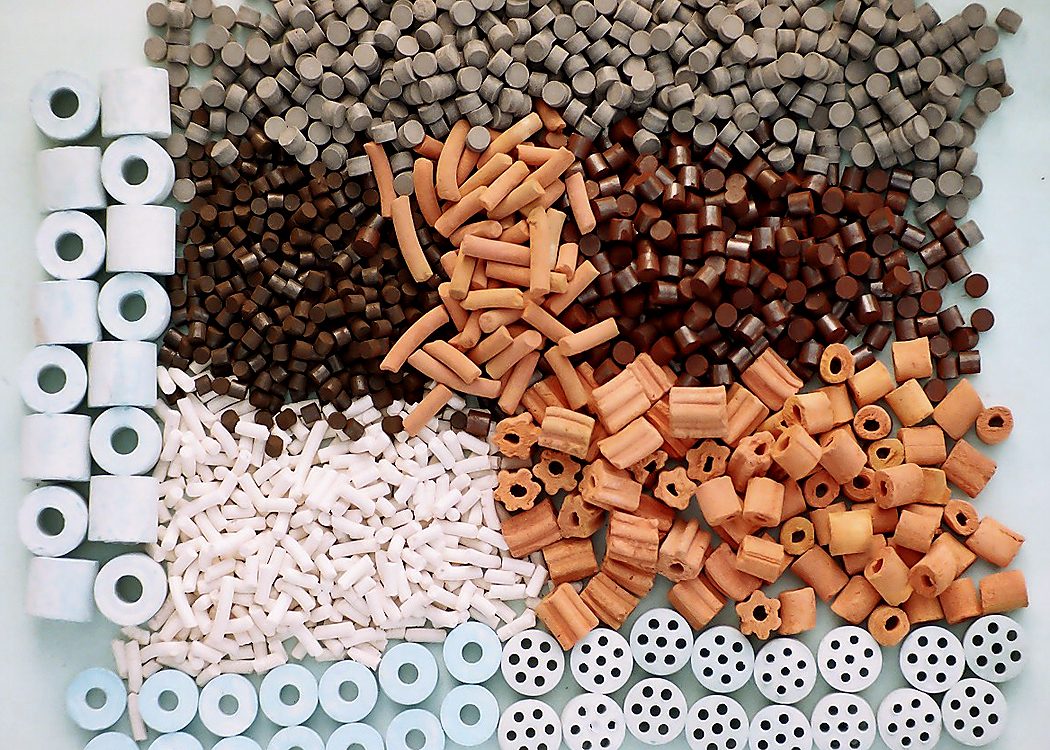 A tray holding many small ceramic catalyst pieces in different shapes: solid cylinders, rings, ribbed rods and discs with holes