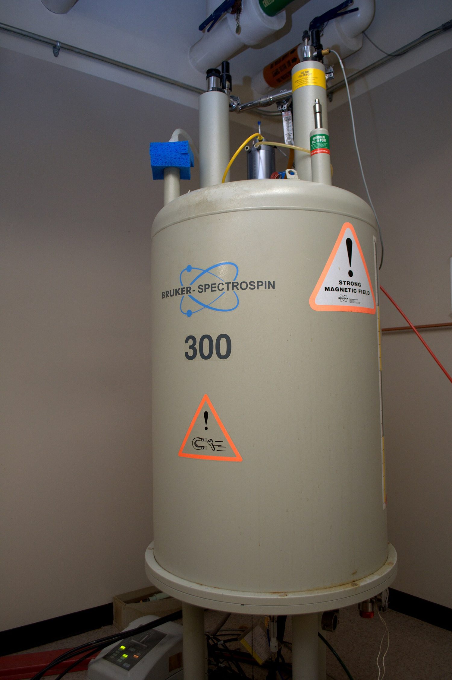 A tall cylindrical NMR spectrometer standing in a laboratory, marked with a strong magnetic field warning
