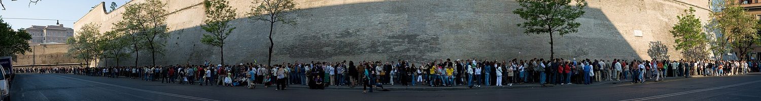 A very long line of people waiting one behind another, stretching along a wall into the distance