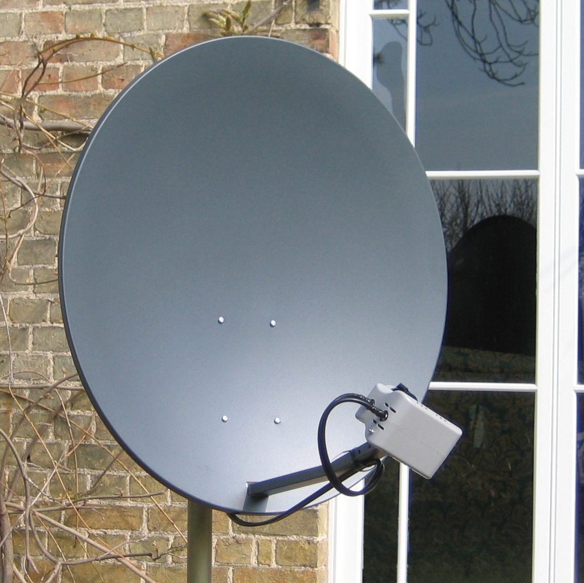 A round grey home satellite dish mounted on a house wall beside a window, with its feed arm reaching out to the front
