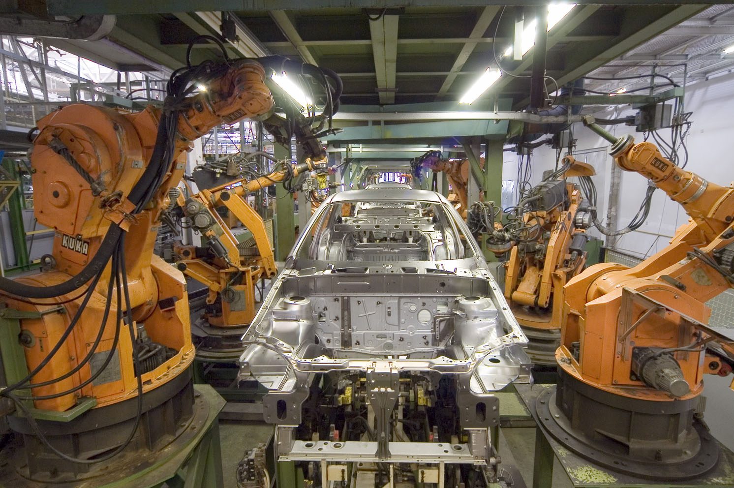 Several orange industrial robot arms welding a car body as it moves along a factory production line