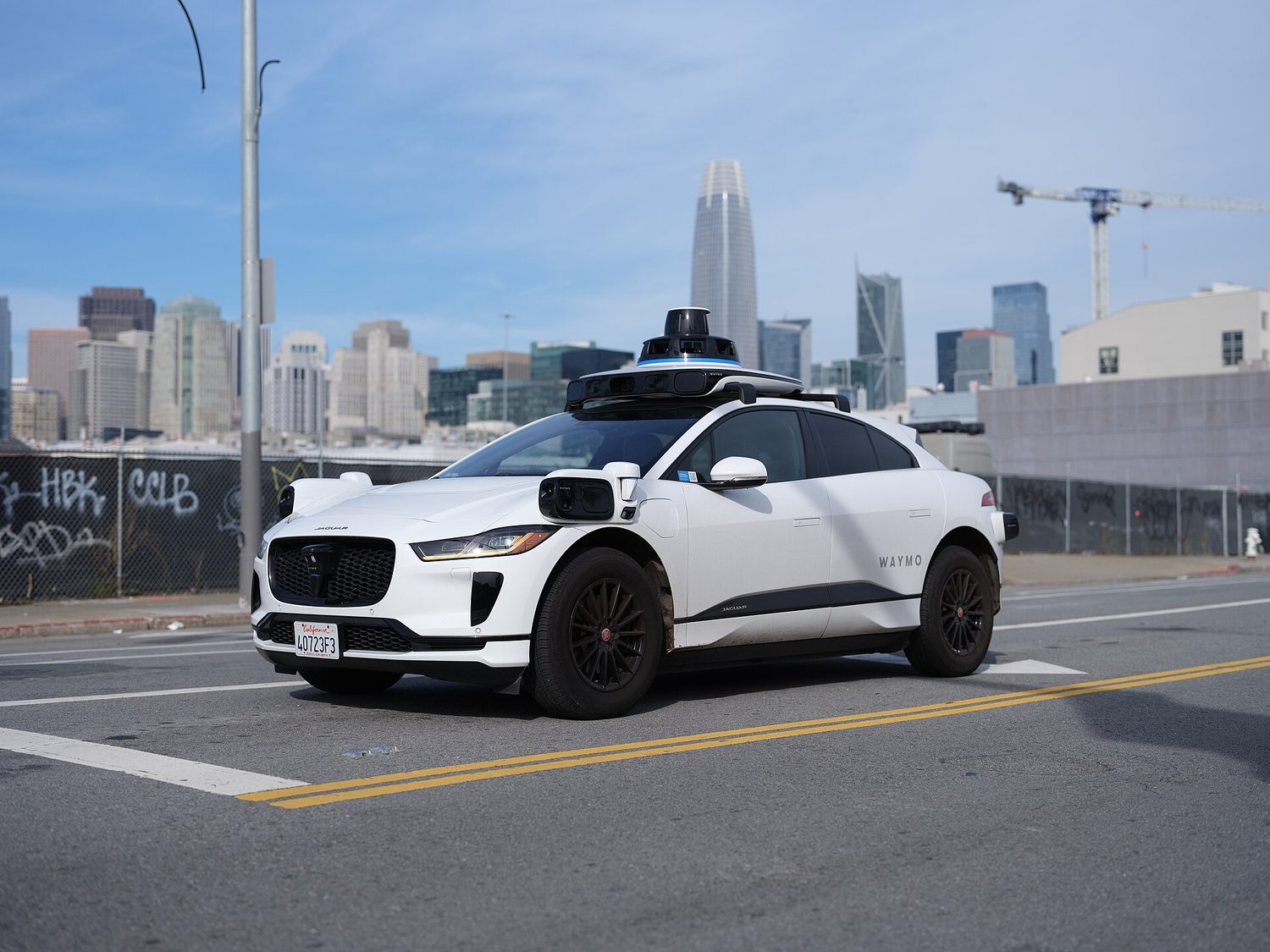 A white Waymo self-driving car on a city street, with a spinning lidar sensor unit on its roof and extra cameras at the front corners