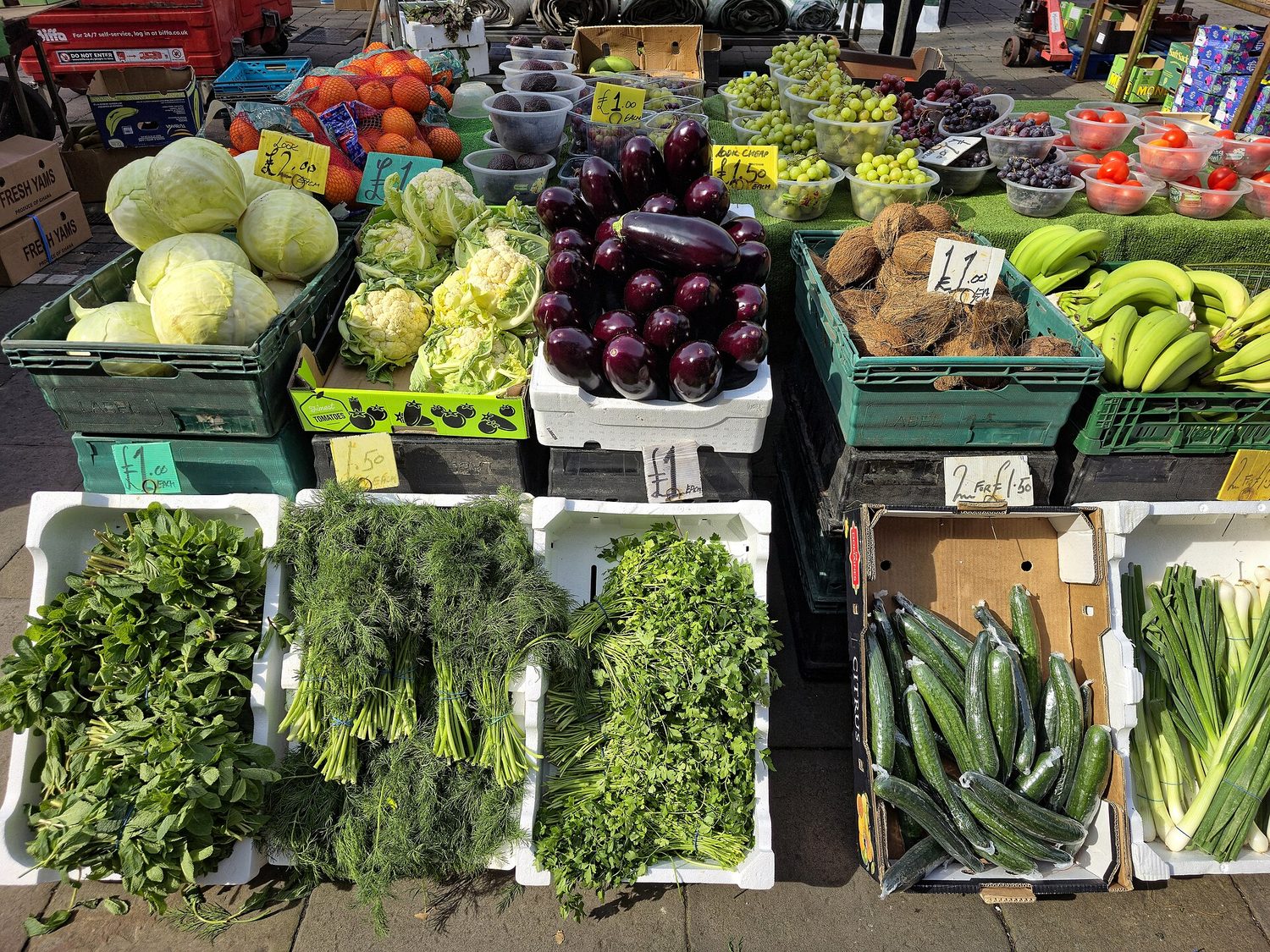 A real outdoor market stall: a greengrocer's vegetable display with handwritten price tags, and shoppers with trolleys choosing produce