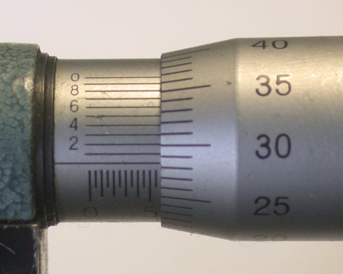 A close-up photograph of a real micrometer scale: the sleeve's main scale on the left meets the rotating thimble scale on the right at the datum line