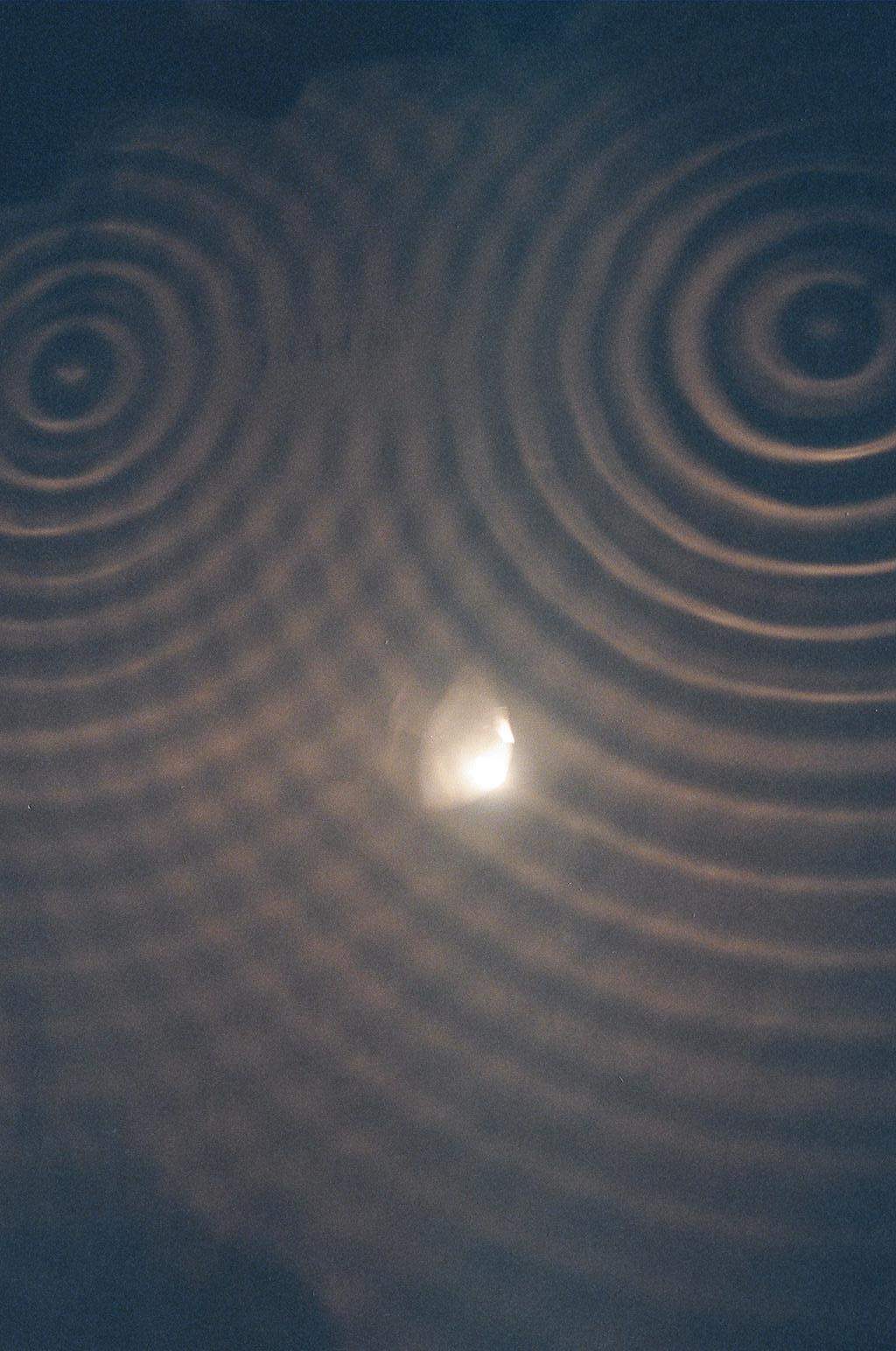 A real ripple-tank photograph: two sets of circular water waves from two side-by-side sources overlap, leaving calm lines (cancellation) fanning out between the two bright sets of ripples