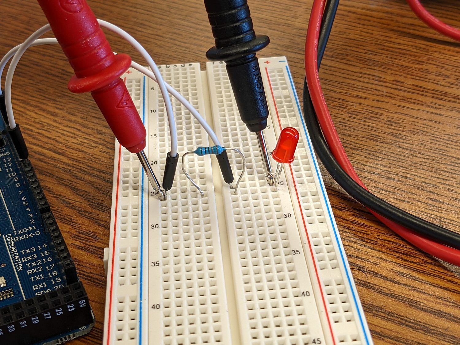A small circuit on a breadboard: a resistor and a glowing red LED are plugged into the rows, wired to a microcontroller, while the red and black probes of a multimeter touch the circuit to take a reading