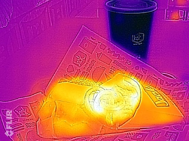A thermal-camera (infrared) image of fast food: a paper bag of fries glowing bright yellow and orange next to a drinks cup that appears dark, all on a purple background