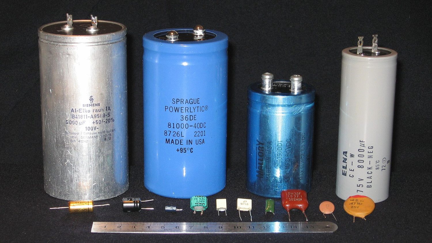 Several real capacitors of very different sizes: four large metal electrolytic cans standing upright, and a row of much smaller film and ceramic capacitors below, with a ruler for scale