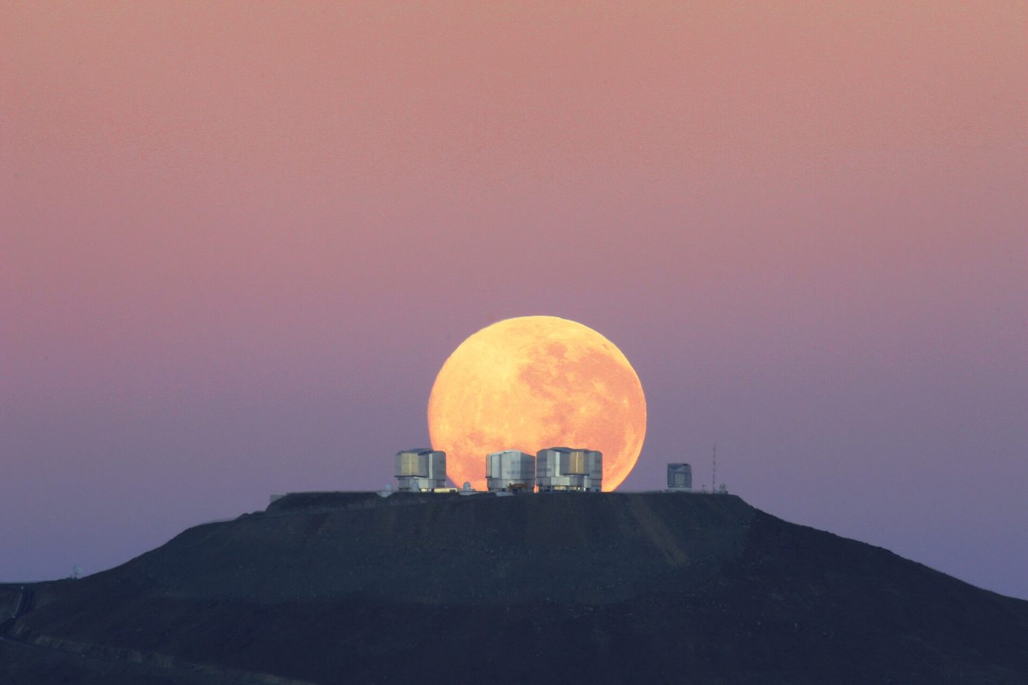 Three large telescope domes silhouetted on a mountain ridge at dusk against a pink sky, with a huge orange full Moon rising directly behind them
