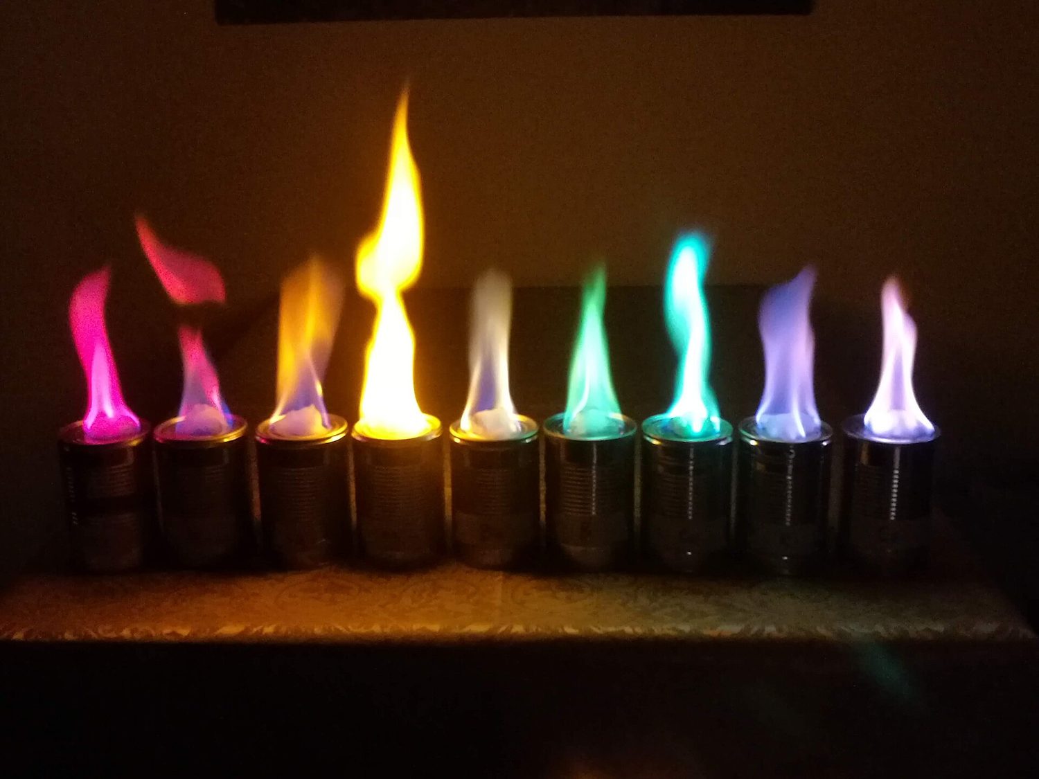 A row of nine burning dishes, each flame a different vivid colour — crimson, orange, yellow, green, blue, violet