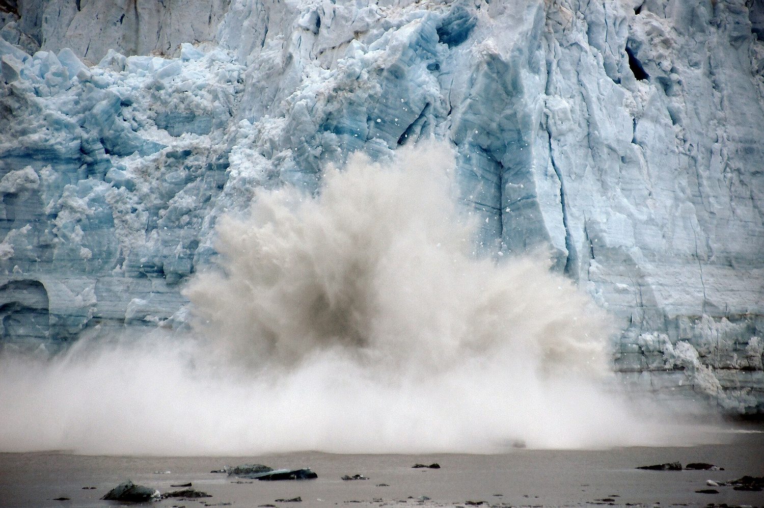 A tall wall of glacier ice with a large chunk crashing into the sea in a cloud of spray