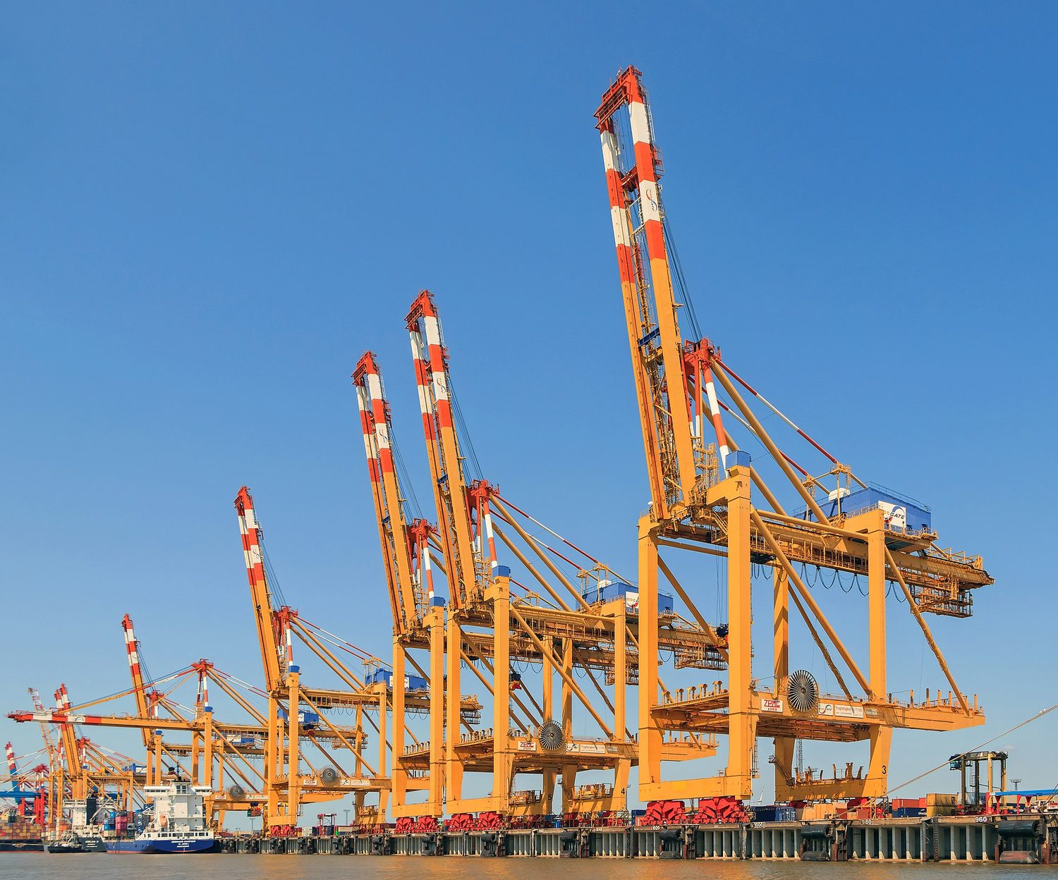 Tall orange ship-to-shore container cranes at a port against a blue sky, with a ship and stacked containers