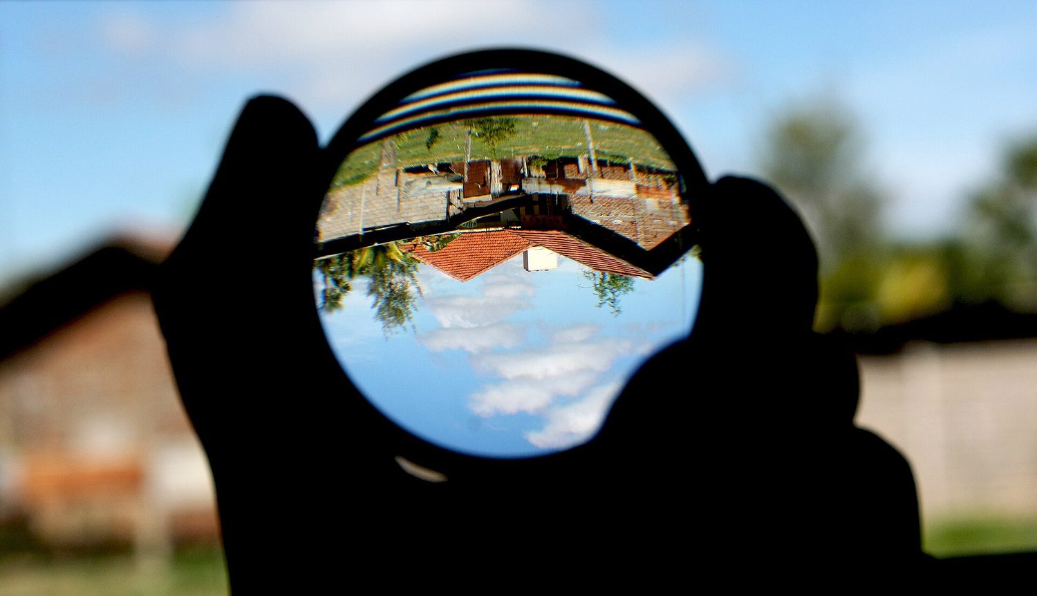A hand holding a round convex lens, through which the houses behind appear upside down