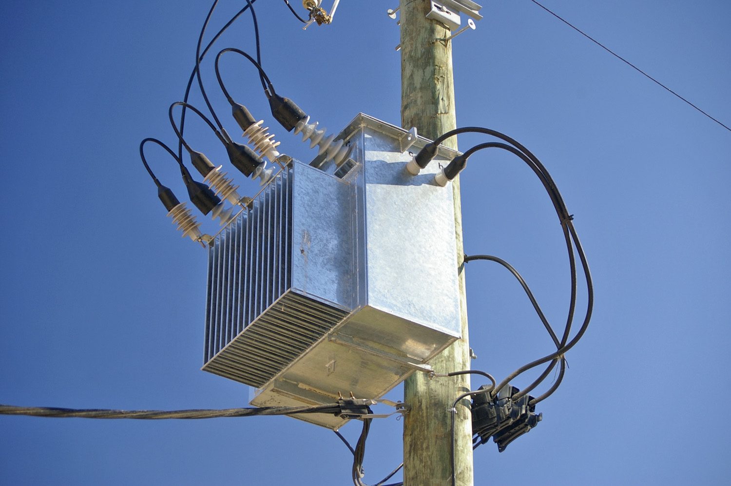 A grey metal transformer fixed to a wooden electricity pole, with thick insulated cables running to it